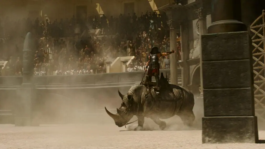 A beefy gladiator rides on top of a huge armored rhinoceros in the middle of the Colosseum as packed crowds watch from the stands in the legacy sequel GLADIATOR II.  