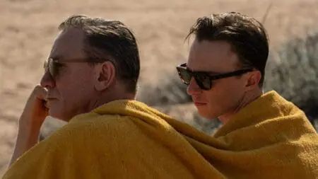 Daniel Craig and Drew Starkey sit together on a South American beach both wearing their sunglasses and sharing a large yellow towel in the movie QUEER.