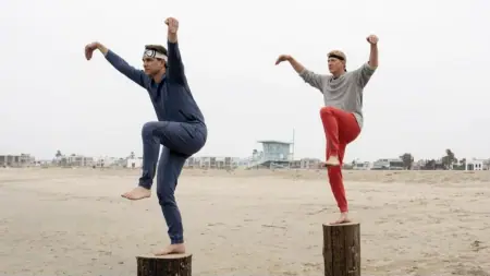 Ralph Macchio and William Zabka stand on two separate wooden stumps on an empty beach practicing the iconic crane kick from the Karate Kid franchise in COBRA KAI Season 6 Part 3, which is included in the complete Netflix February 2025 list of new arrivals.