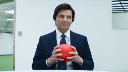 Lumon Industries employee and severed worker Mark Scout, played by Adam Scott, gives off a huge smile while holding a small bright red rubber ball in SEVERANCE Season 2 streaming only on Apple TV+.
