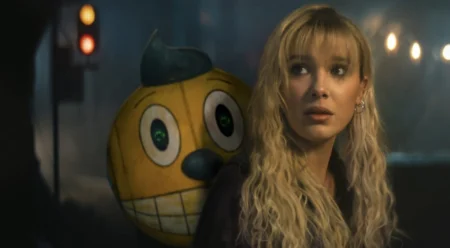 Actress Millie Bobby Brown sporting curly blonde hair stars as the young woman Michelle accompanied by her small sentient toy robot companion with large cartoon eyes and a smiley face painted on its round yellow head in the sci-fi adventure movie THE ELECTRIC STATE, which is featured in the Netflix March 2025 list of new arrivals.