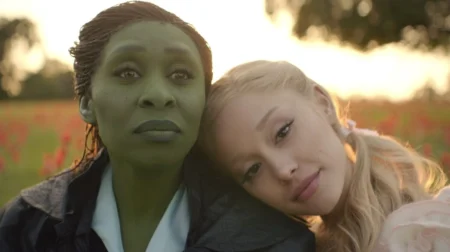 Ariana Grande as Glinda Upland rests her head on the shoulder of the green witch Elphaba Thropp, played by Cynthia Erivo, as the two sit together on an open field in front of a beautiful sunset in the movie adaptation of WICKED, which is featured in the Peacock March 2025 list of new arrivals.