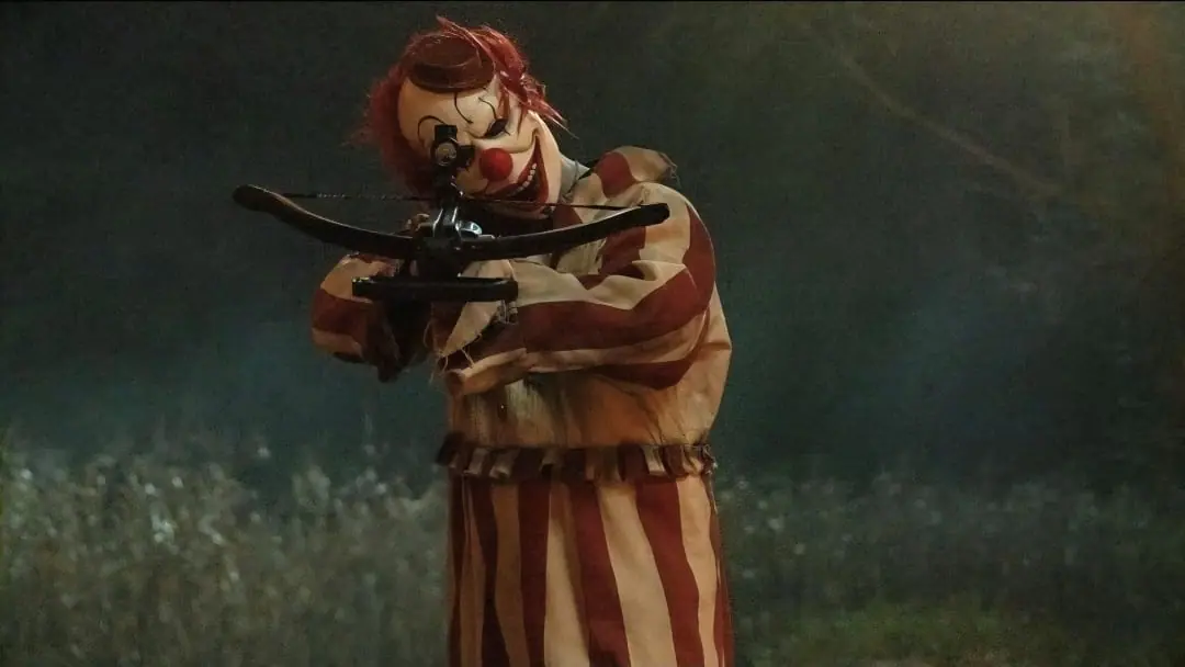 Frendo the Clown aims a large crossbow at the camera in a horrifying shot from the 2025 meta slasher film CLOWN IN A CORNFIELD. 