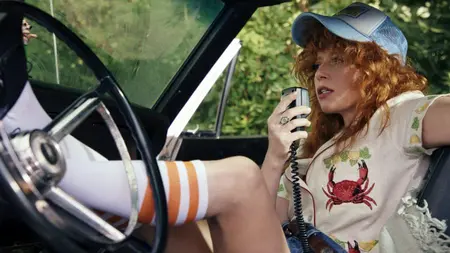 Natasha Lyonne stars as former cocktail waitress Charlie Cale laying back in a muscle car with her legs up on the steering wheel as she speaks into a handheld radio microphone in POKER FACE Season 2 streaming only on Peacock.