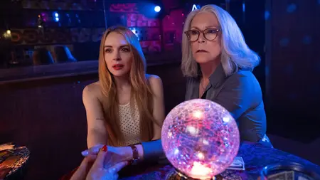 Lindsay Lohan and Jamie Lee Curtis look confused and worried as they sit next to each other at a fortune teller's table with a large, shiny, and purple magic ball on top in the Disney legacy sequel FREAKIER FRIDAY.