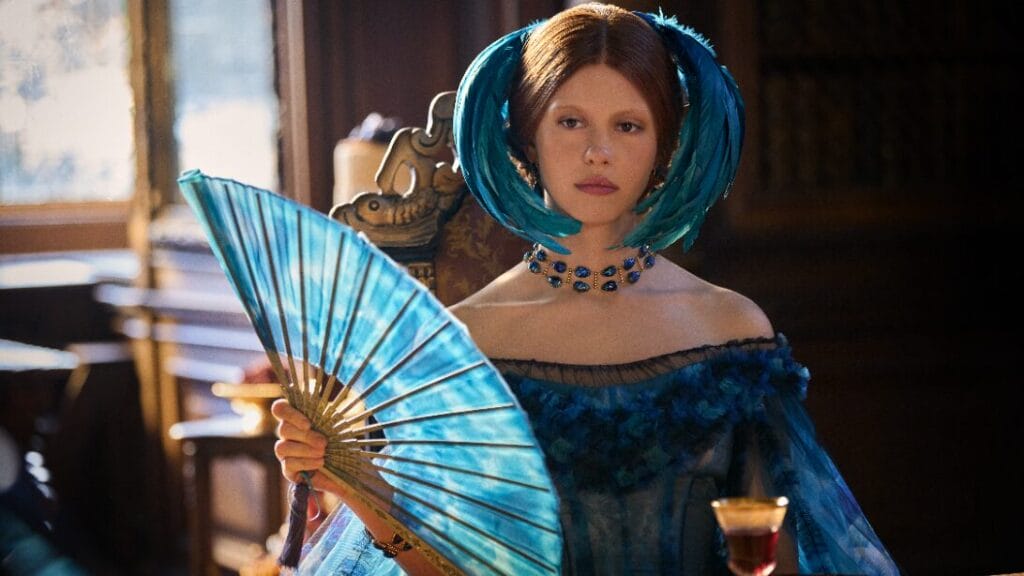Mia Goth stars as  Elizabeth Lavenza wearing a lavish 19th-century blue dress and head piece as she cools herself with a large matching blue folding fan in Guillermo del Toro's FRANKENSTEIN. 
