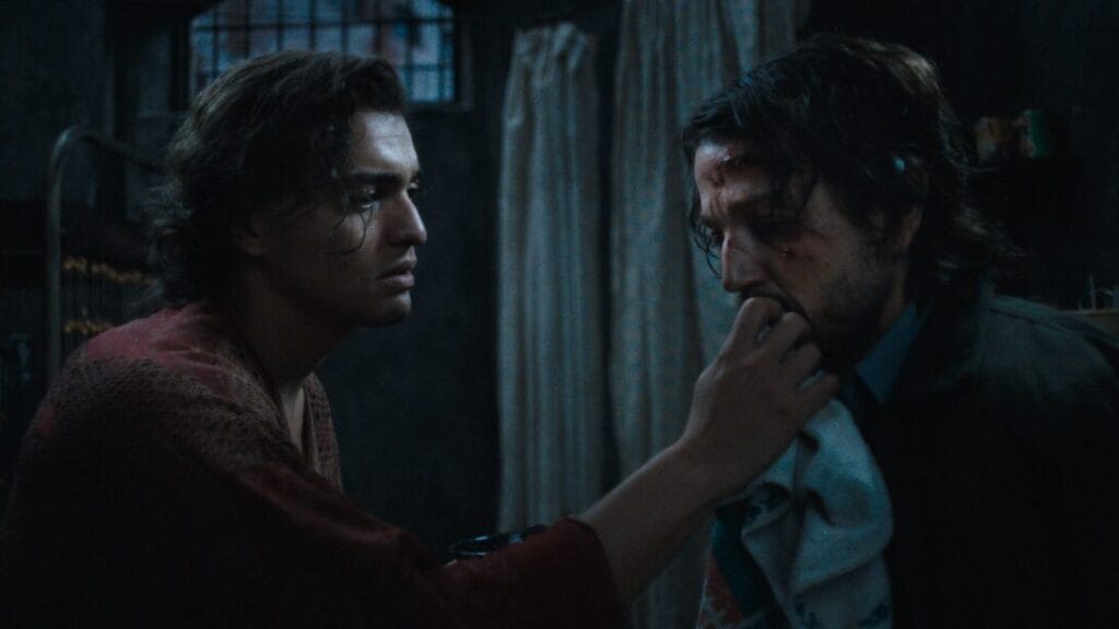 Tonatiuh holds up Diego Luna's injured face inside a grimy jail cell as the two portray political prisoners in 1983 Argentina in the movie musical adaptation of KISS OF THE SPIDER WOMAN.