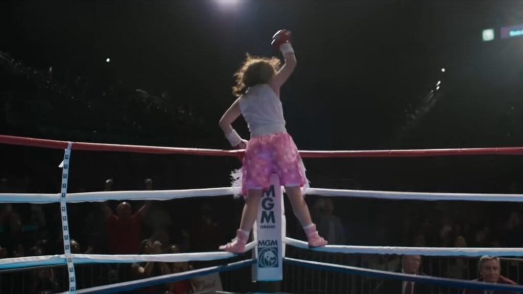 Sydney Sweeney stars as famous female boxer Christy Martin jumping onto the ropes of the boxing ring while raising her glove in the air with pure happiness after winning a match in the CHRISTY biopic. 