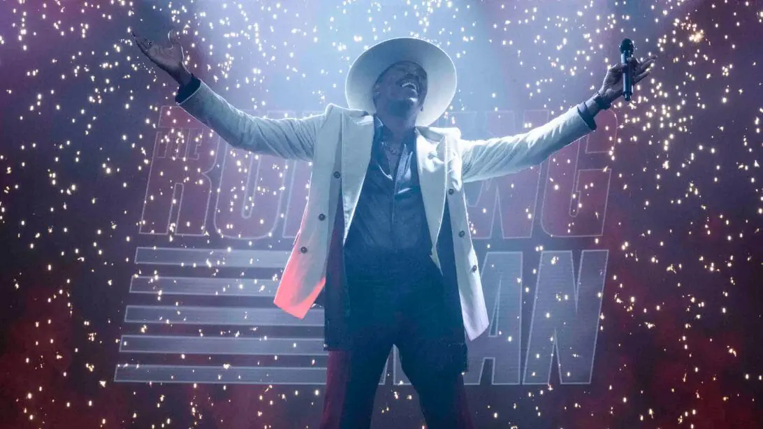 Colman Domingo stuns as TV host Bobby T. in a white suit and fedora while he stands in the middle of The Running Man stage as red and white confetti falls all around him. 