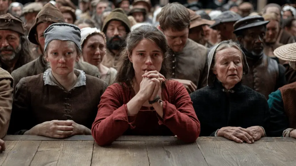Jessie Buckley stars as Agnes Shakespeare holding up her hands in a praying position as she leans onto a large wooden outdoor theatre stage in front of a packed crowd of spectators in director Chloé Zhao's HAMNET.