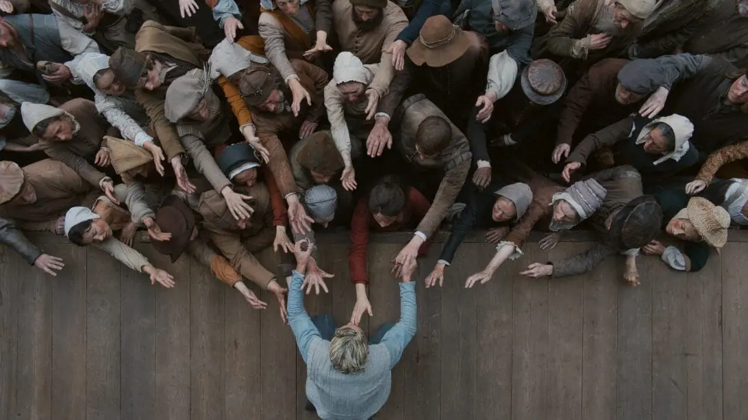 A majestic overhead shot of Noah Jupe playing Hamlet on the large wooden stage of the Globe Theatre in Elizabethan London, while he bows down and reaches his arms out to a large audience who are all trying to hold him back, in the movie adaptation of HAMNET.