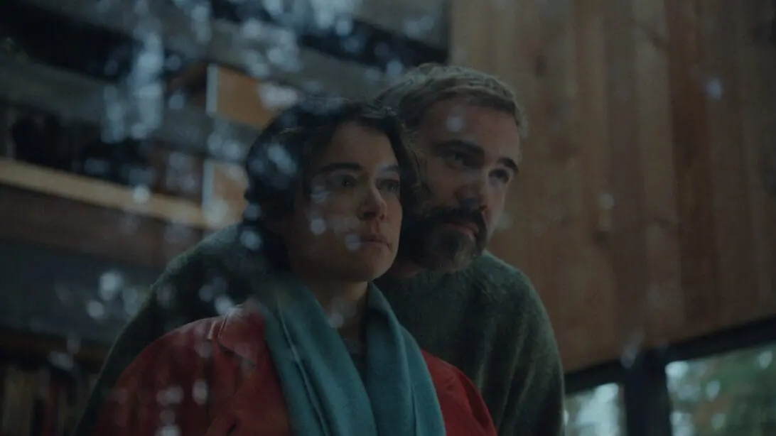 Rossif Sutherland embraces Tatiana Maslany with a romantic hug from behind as the couple stare out of a large glass window in their remote house in the woods in the horror movie KEEPER.