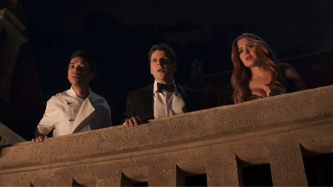 Justice Smith, Jesse Eisenberg, and Isla Fisher look down at a fancy party from a dark rooftop while wearing luxurious suits and dresses in NOW YOU SEE ME 3. 