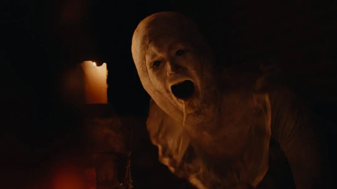 A horrifying pale-faced humanoid monster with wax dripping out of its mouth crawls next to a lit candle in a dark room in KEEPER directed by Osgood Perkins. 