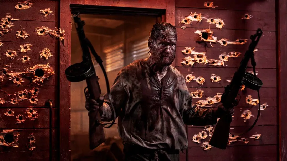 Jorma Tommila as the legendary Finnish commando Aatami Korpi poses with two large machine guns in front of a red barn covered in insanely large bullet holes in SISU 2. 