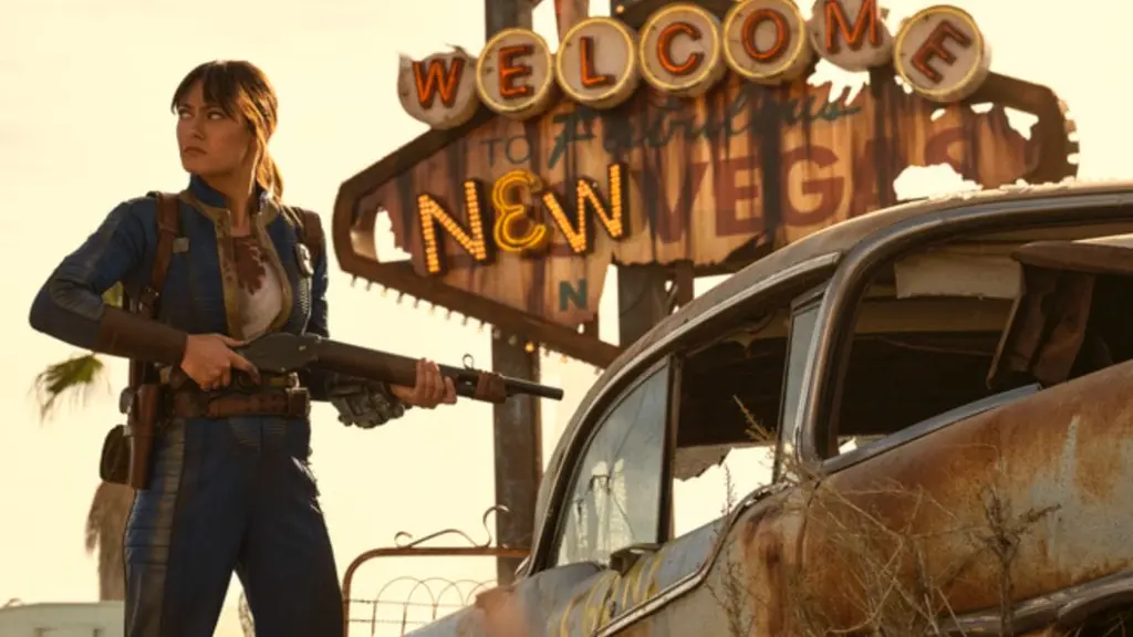 Ella Purnell stars as vault dweller Lucy in FALLOUT Season 2, standing in front of the iconic New Vegas neon sign from the games while holding a large rifle.