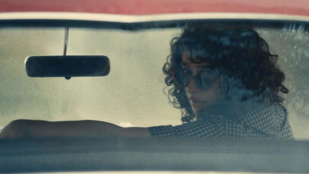 Odessa A'zion stars as the young and reckless Rachel Mizler looking back from the front seat of an old car with her sunglasses down in the A24 sports drama MARTY SUPREME.