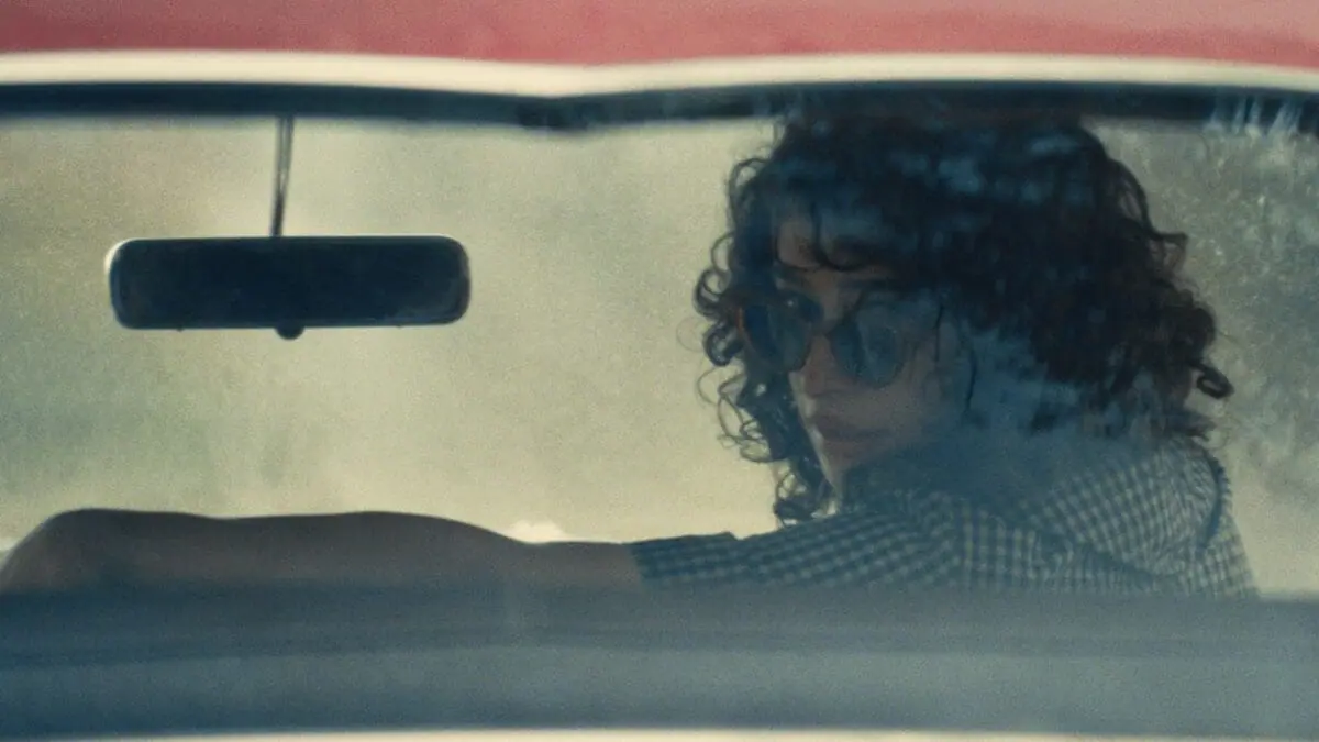 Odessa A'zion stars as the young and reckless Rachel Mizler looking back from the front seat of an old car with her sunglasses down in the A24 sports drama MARTY SUPREME. 