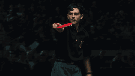 Timothée Chalamet stars as star ping pong player Marty Mauser raising his paddle as he points at his opponent across the table with a fierce look of determination in the A24 sports drama MARTY SUPREME.
