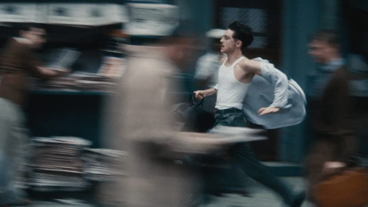 A blurry shot of Timothée Chalamet running down a busy street in 1950s New York City with his shirt almost flying off in the A24 film MARTY SUPREME. 