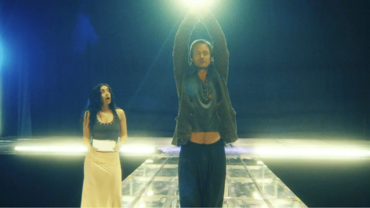 Alexander Skarsgård strikes a hilarious dance pose next to pop star Charli xcx on a large concert stage during rehearsal for her brat tour in the A24 mockumentary THE MOMENT. 