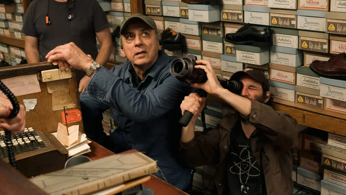 Cinematographer Darius Khondji sits next to director Josh Safdie who is holding up a small camera to his eye as the two block a scene inside a shoe store with '50s time period props on the set of the A24 movie MARTY SUPREME.