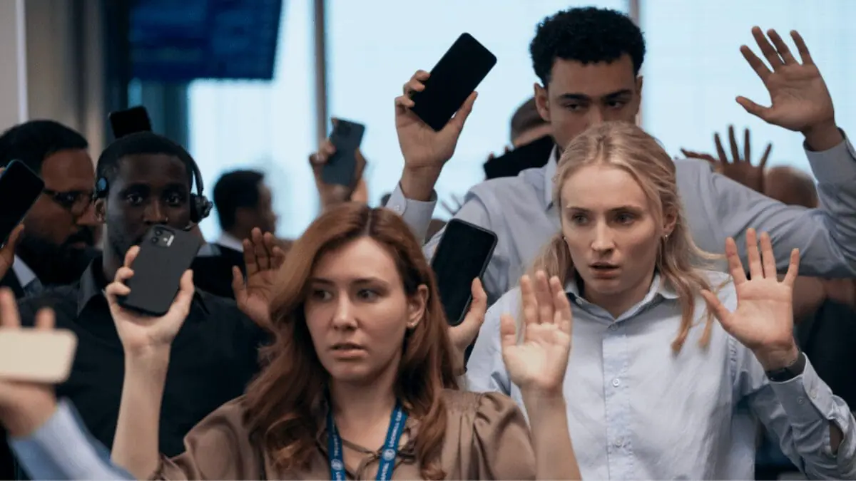 Sophie Turner and Archie Madekwe raise their hands in fear and turn in their smart phones as their pension fund investment company gets taken hostage by armed thieves in the new Prime Video original series STEAL, which is premiering in January 2026.