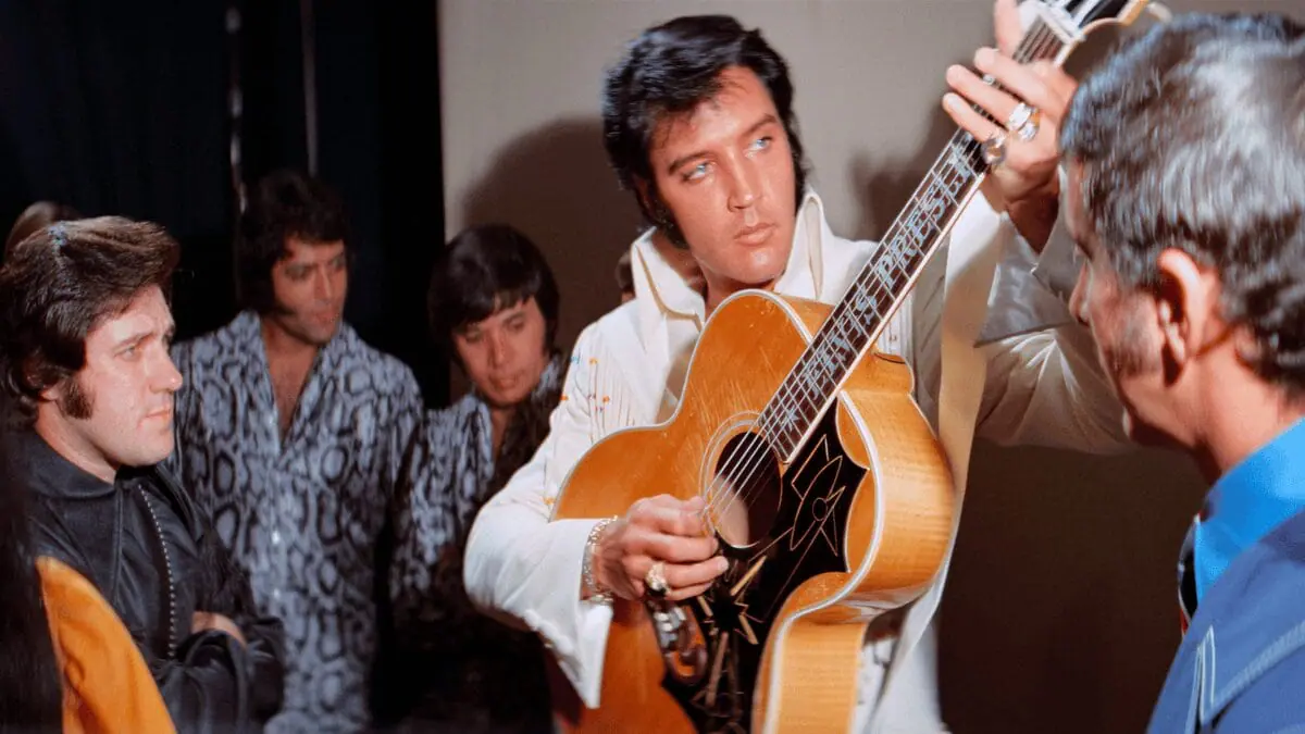 A rare behind-the-scenes image of Elvis Presley tuning his guitar backstage while wearing one of his iconic white leather suits in Baz Luhrmann's music documentary EPiC: ELVIS PRESLEY IN CONCERT.