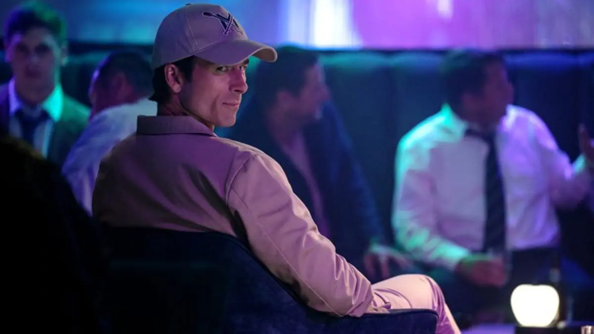 Glen Powell looks back towards the camera and gives a small devious smile while doing some incognito spying at a bar full of young business men in the social class thriller HOW TO MAKE A KILLING. 