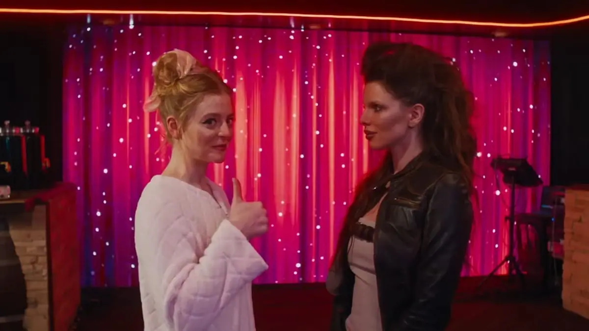 Anna Baryshnikov looks to the camera and gives a thumbs up while being interviewed by Julia Fox as part of a fashion competition show in the satirical comedy IDIOTKA. 