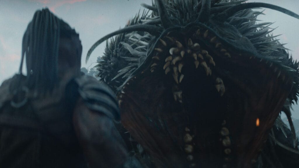 The giant and mighty unkillable Kalisk beast roars in front of the small Yautja warrior Dek and shows off its many rows of sharp teeth in the epic sci-fi blockbuster PREDATOR: BADLANDS. 
