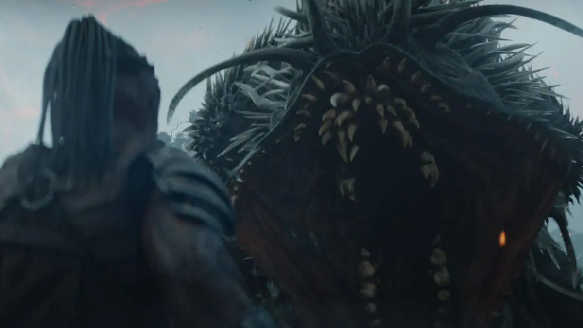 The giant and mighty unkillable Kalisk beast roars in front of the small Yautja warrior Dek and shows off its many rows of sharp teeth in the epic sci-fi blockbuster PREDATOR: BADLANDS. 