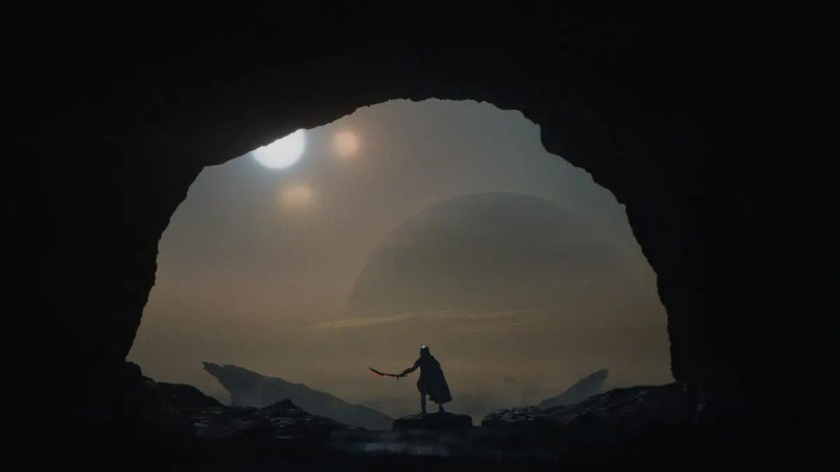 An epic wide shot of a young Yautja warrior taking out his large laser sword as he stands at the entrance of a giant cave on an alien planet with multiple moons and suns seen behind him in the distance in the sci-fi blockbuster PREDATOR: BADLANDS.