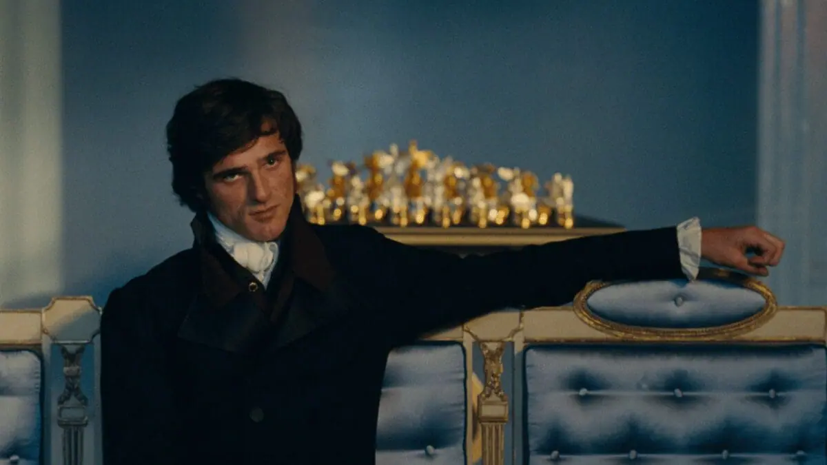 Jacob Elordi stars as Heathcliff sitting down in a luxurious couch while wearing 18th-century classic attire in Emerald Fennell's film adaptation of WUTHERING HEIGHTS.