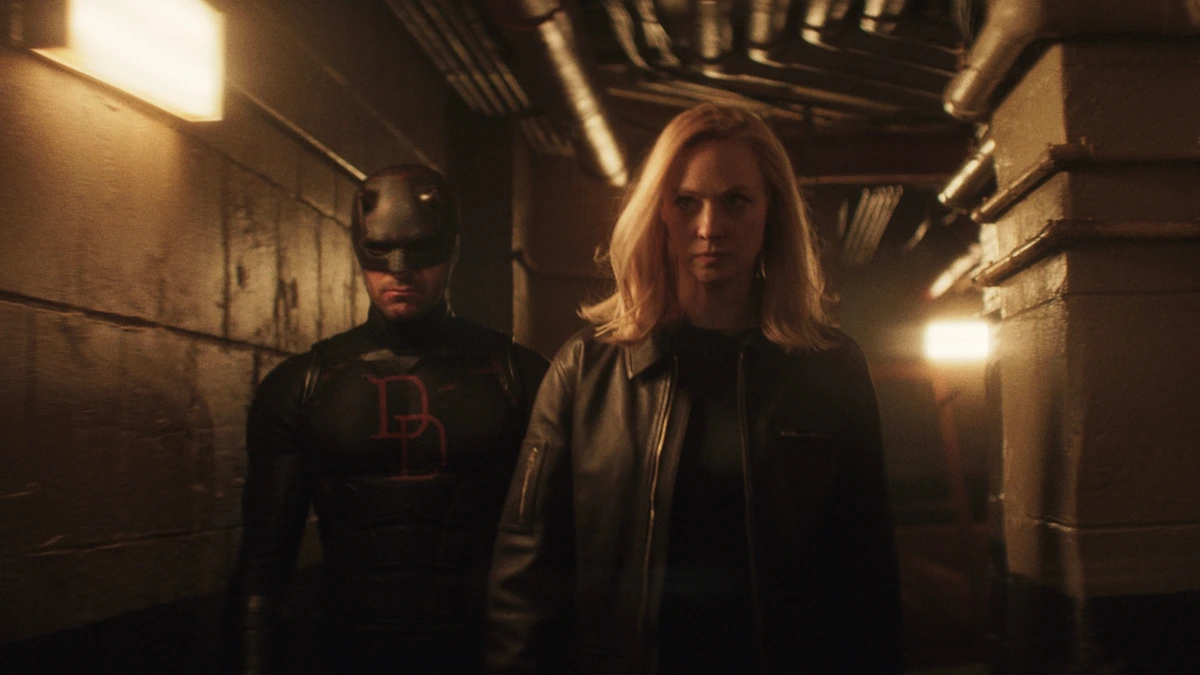 Daredevil wearing his new black suit walks alongside Karen Page down a dimly-lit industrial hallway in DAREDEVIL: BORN AGAIN Season 2.