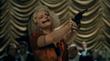 Jessie Buckley stars as the Bride of Frankenstein aiming a pistol in the air as she holds a bunch of people in a fancy ballroom hostage in Maggie Gyllenhaal's THE BRIDE!