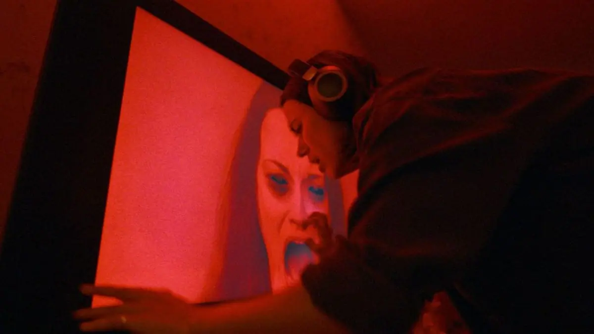 Nina Kiri panics as she tries to turn off a large TV with a bright red image of a scary looking woman screaming in the horror film UNDERTONE. 