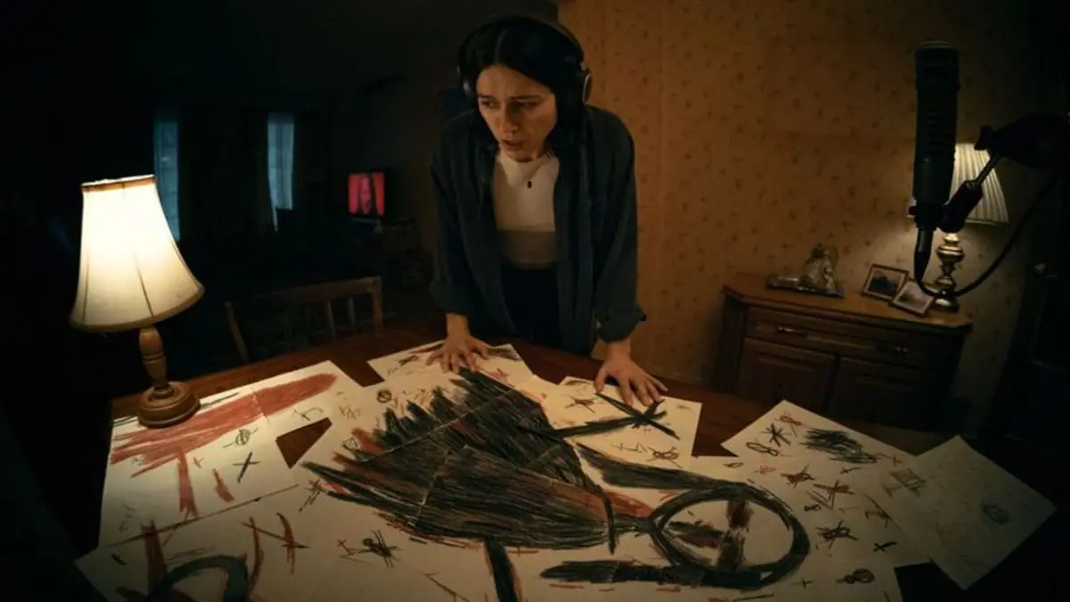 Nina Kiri looks down at a large table covered with creepy looking black drawings of a demonic figure in the horror movie UNDERTONE. 