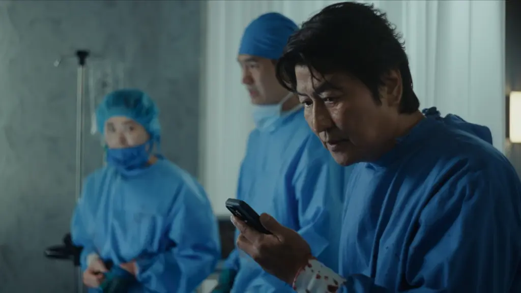 Esteemed Korean actor Song Kang-ho looks at his phone with a shocked facial expression while wearing blue scrubs as Dr. Kim the plastic surgeon in BEEF Season 2. 