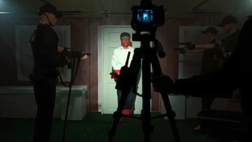 A man gagged with his hands tied stands frozen in fear in front of a tripod camera while surrounded by mannequins in police uniforms in the 2026 horror remake of FACES OF DEATH. 