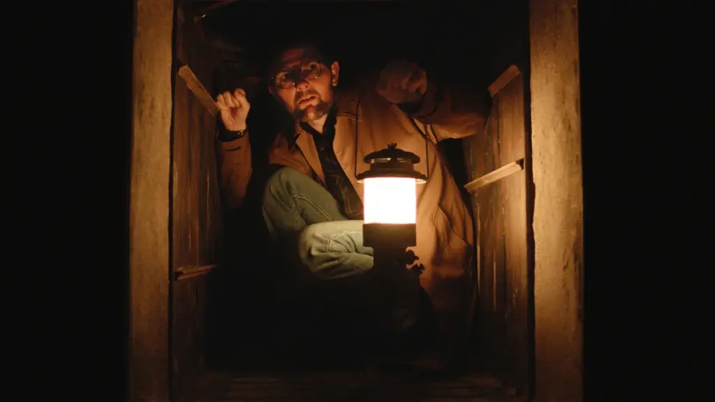 Adam Scott kneels down as he squeezes through a small and dark square opening while holding an old gas lamp in the horror movie HOKUM. 