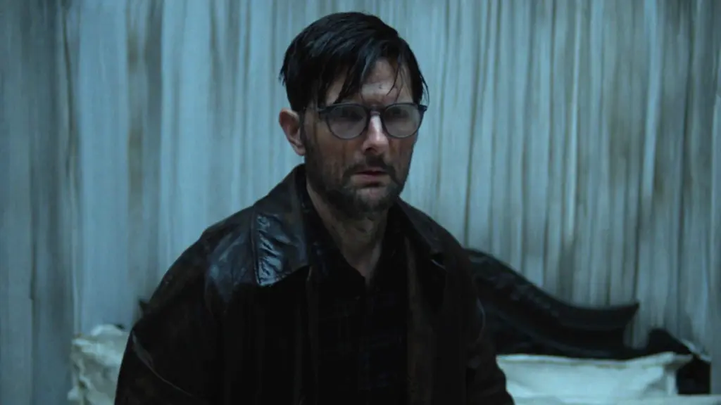 Adam Scott makes a shocked face while his eye glasses and whole body is covered in mud and dirt in the Irish folk horror film HOKUM.
