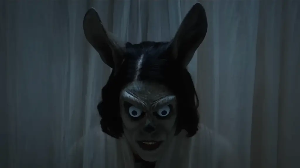 A person wearing an extremely creepy humanoid rabbit mask sticks their head through a curtain in the Irish folk horror movie HOKUM.