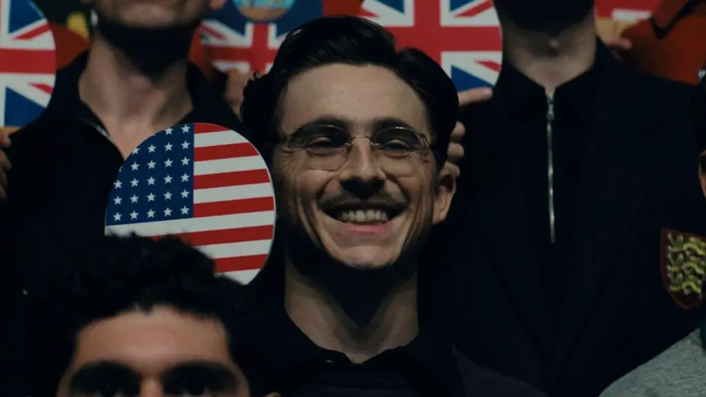 A close up shot of Timothée Chalamet smiling while holding up a ping pong paddle with the American flag printed on it for a tournament group photo in A24's MARTY SUPREME, which is included in the HBO Max April 2026 list of new movies and TV shows.