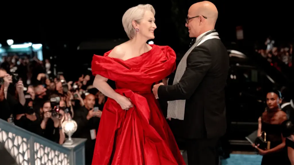 Meryl Streep looks stunning in a lavish red dress next to Stanley Tucci  wearing a sleek black tuxedo and gray scarf as the two walk down a red carpet surrounded by paparazzi and flashing camera lights in THE DEVIL WEARS PRADA 2. 