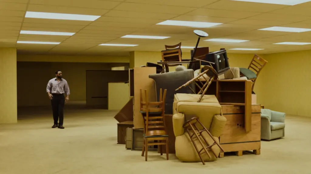 Chiwetel Ejiofor walks next to a large pile of chairs, ottomans, sofas, and office desks inside the endless dimension of the Backrooms in the A24 film. 