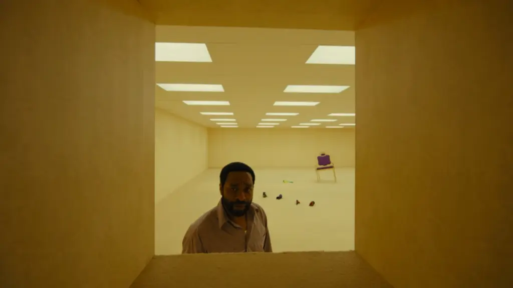 Chiwetel Ejiofor looks into a large box-shaped crawl space in the empty, never-ending office spaces of the Backrooms in the A24 film. 