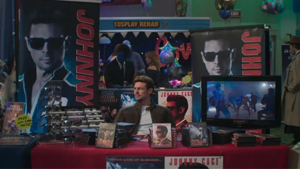 Johnny Cage played by Karl Urban sits lonely at his fan table at a convention showing off all his famous action films in MORTAL KOMBAT II. 