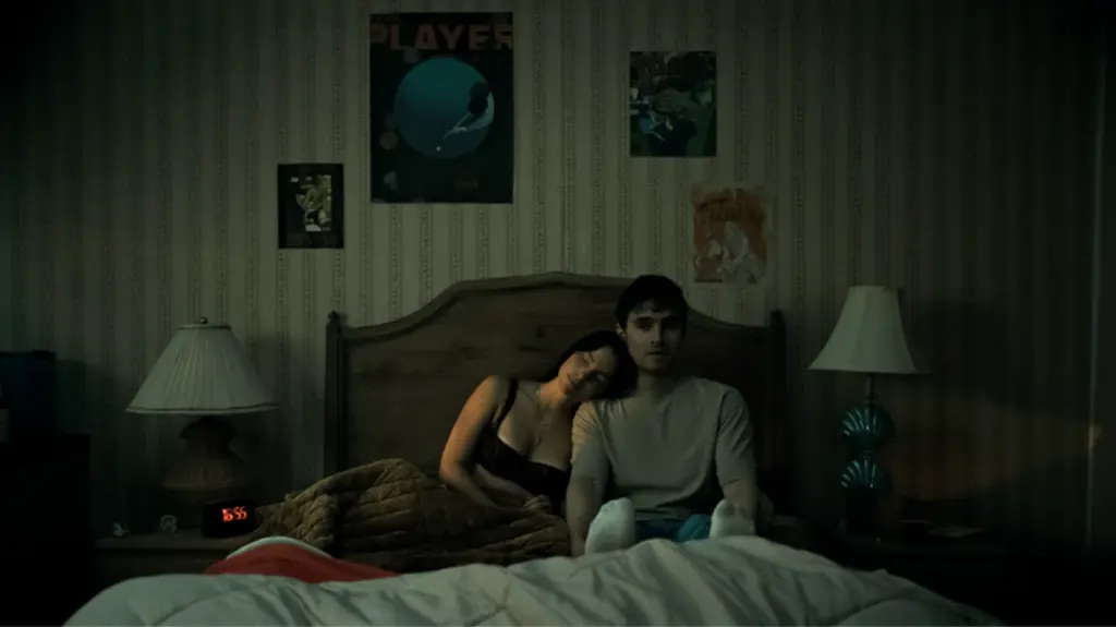A creepy shot of the boyfriend protagonist Bear sitting up in his dark bedroom while his insanely obsessed girlfriend Nikki lays on his shoulder in the horror movie OBSESSION. 