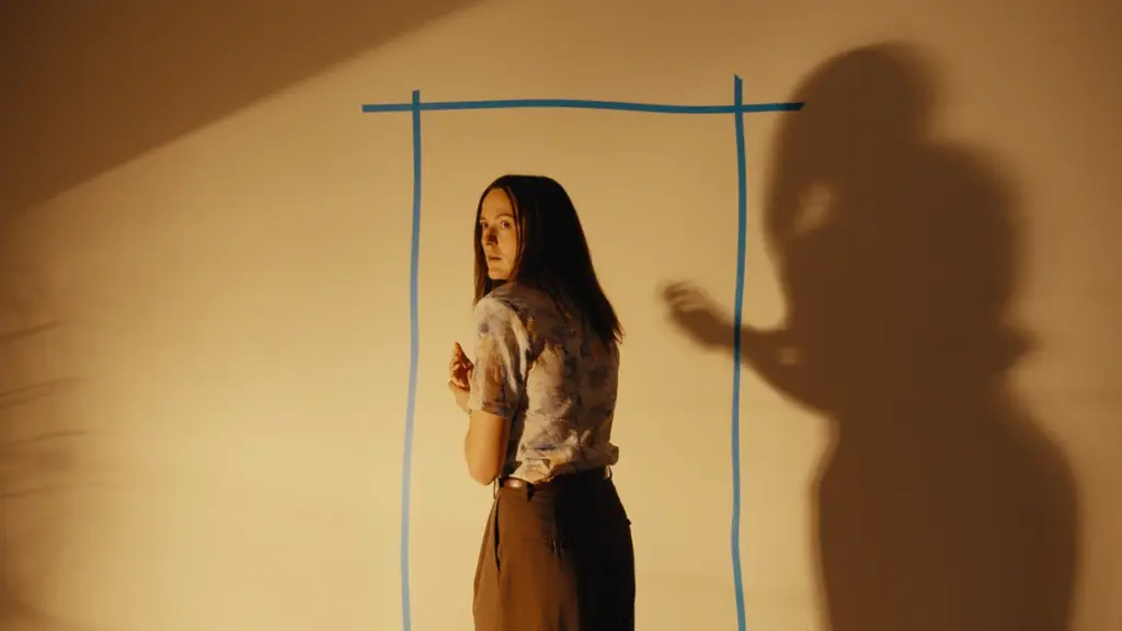 Renate Reinsve looks back in fear as she prepares to enter the Backrooms through a portal drawn out as a square door on an empty wall with blue marker.